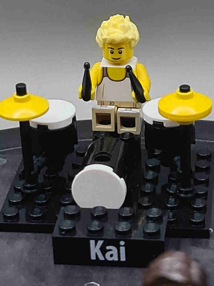 Kai is represented by a Lego figure in classic Lego yellow. Kai is playing a Lego drumkit.
